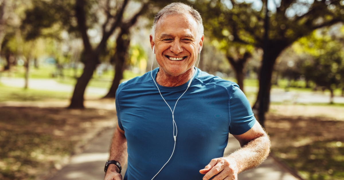 Smiling middle-aged man jogging outdoors in a park, wearing blue athletic shirt and earphones