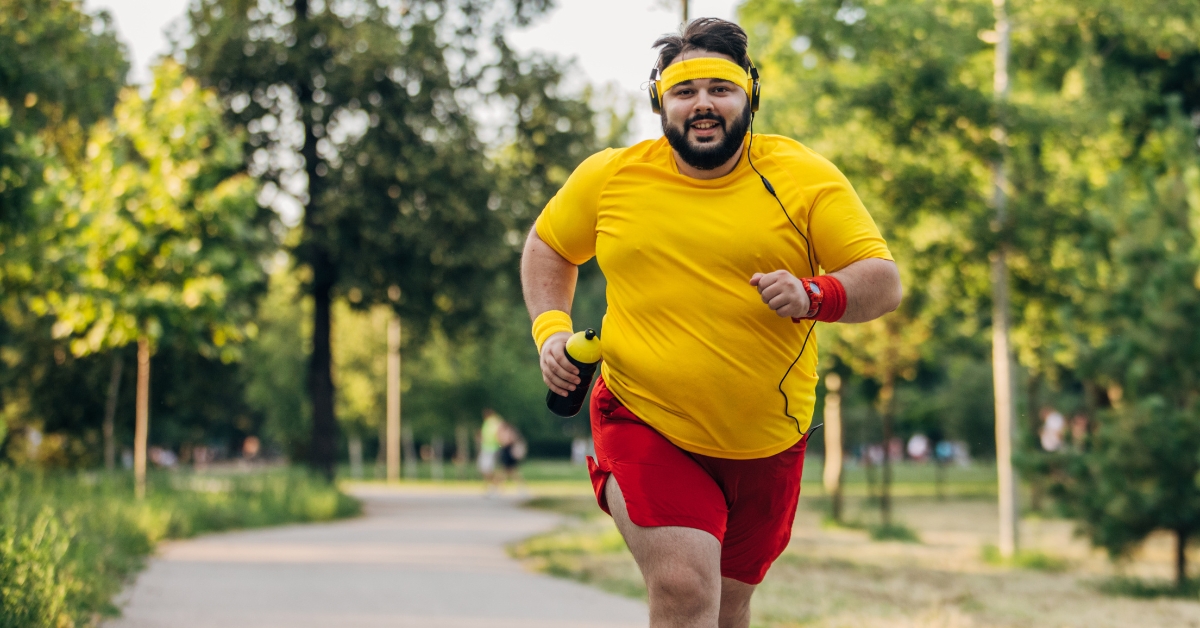 Overweight man jogging in a park wearing yellow shirt, red shorts, and headphones
